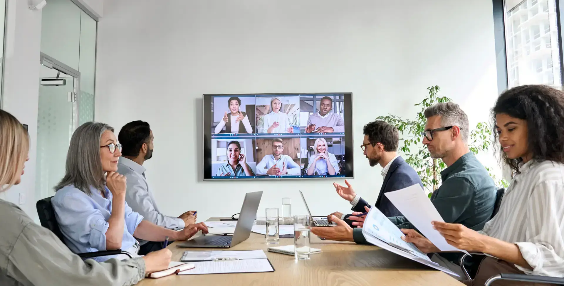 Ein Team sitzt am Konferenztisch, weitere Mitarbeiter sind auf einem Bildschirm zu sehen und arbeiten an einem ERP-System.